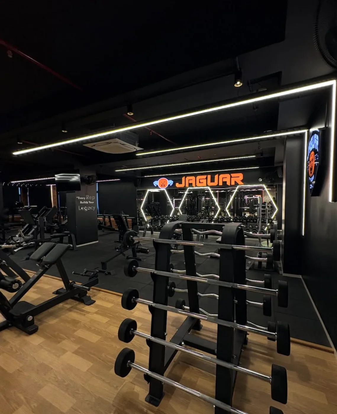 Barbell rack and benches on the Jaguar Fitness Den training floor, with the illuminated orange JAGUAR sign and hexagon mirror lights behind